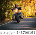 Low angle action photograph of a motorcyclist in black protective gear leaning into a curve on an asphalt road surrounded by golden autumn trees, with warm sunlight and motion blur. 143700161