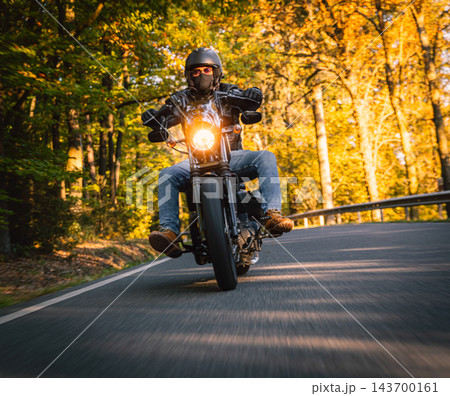 Low angle action photograph of a motorcyclist in black protective gear leaning into a curve on an asphalt road surrounded by golden autumn trees, with warm sunlight and motion blur. Low angle action photograph of a motorcyclist in black protective gear leaning into a curve on an asphalt road surrounded by golden autumn trees, with warm sunlight and motion blur. 143700161
