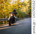 Low angle action photograph of a motorcyclist in black protective gear leaning into a curve on an asphalt road surrounded by golden autumn trees, with warm sunlight and motion blur. 143700162