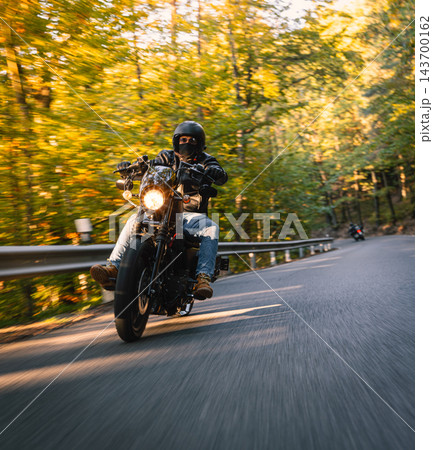 Low angle action photograph of a motorcyclist in black protective gear leaning into a curve on an asphalt road surrounded by golden autumn trees, with warm sunlight and motion blur. Low angle action photograph of a motorcyclist in black protective gear leaning into a curve on an asphalt road surrounded by golden autumn trees, with warm sunlight and motion blur. 143700162
