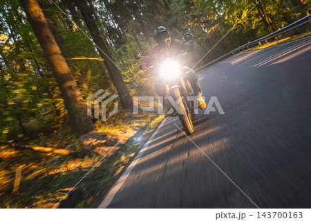 Motorcyclist wearing a black helmet and dark riding gear approaching the camera on a winding asphalt road through a sunlit green and golden autumn forest, with dynamic motion blur. Motorcyclist wearing a black helmet and dark riding gear approaching the camera on a winding asphalt road through a sunlit green and golden autumn forest, with dynamic motion blur. 143700163