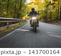 Motorcyclist wearing a black helmet and dark riding gear approaching the camera on a winding asphalt road through a sunlit green and golden autumn forest, with dynamic motion blur. 143700164