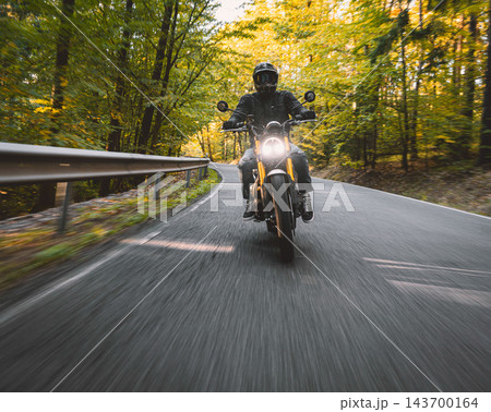 Motorcyclist wearing a black helmet and dark riding gear approaching the camera on a winding asphalt road through a sunlit green and golden autumn forest, with dynamic motion blur. Motorcyclist wearing a black helmet and dark riding gear approaching the camera on a winding asphalt road through a sunlit green and golden autumn forest, with dynamic motion blur. 143700164