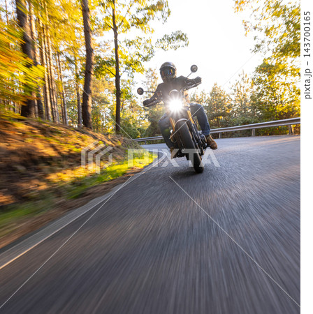 Motorcyclist wearing a black helmet and dark riding gear approaching the camera on a winding asphalt road through a sunlit green and golden autumn forest, with dynamic motion blur. Motorcyclist wearing a black helmet and dark riding gear approaching the camera on a winding asphalt road through a sunlit green and golden autumn forest, with dynamic motion blur. 143700165