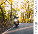 Motorcyclist wearing a black helmet and dark riding gear approaching the camera on a winding asphalt road through a sunlit green and golden autumn forest, with dynamic motion blur. 143700166