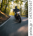 Motorcyclist wearing a black helmet and dark riding gear approaching the camera on a winding asphalt road through a sunlit green and golden autumn forest, with dynamic motion blur. 143700167