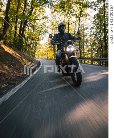 Motorcyclist wearing a black helmet and dark riding gear approaching the camera on a winding asphalt road through a sunlit green and golden autumn forest, with dynamic motion blur. Motorcyclist wearing a black helmet and dark riding gear approaching the camera on a winding asphalt road through a sunlit green and golden autumn forest, with dynamic motion blur. 143700167