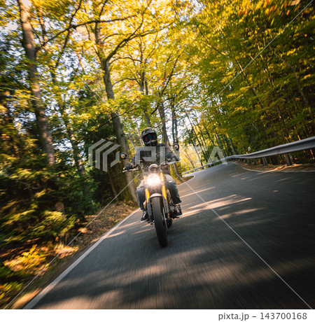 Motorcyclist wearing a black helmet and dark riding gear approaching the camera on a winding asphalt road through a sunlit green and golden autumn forest, with dynamic motion blur. Motorcyclist wearing a black helmet and dark riding gear approaching the camera on a winding asphalt road through a sunlit green and golden autumn forest, with dynamic motion blur. 143700168