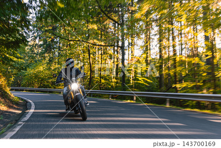 Motorcyclist wearing a black helmet and dark riding gear approaching the camera on a winding asphalt road through a sunlit green and golden autumn forest, with dynamic motion blur. Motorcyclist wearing a black helmet and dark riding gear approaching the camera on a winding asphalt road through a sunlit green and golden autumn forest, with dynamic motion blur. 143700169