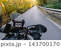 First person POV photograph from a moving motorcycle showing the handlebars, digital dashboard, mirrors and rider's gloved hand while traveling along an asphalt road through a green and golden autumn 143700170