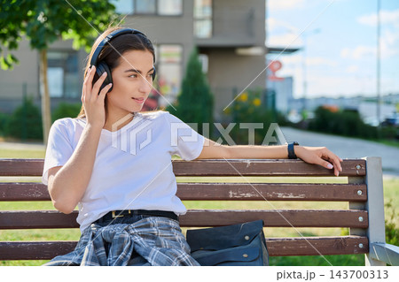 Young woman in headphones listening to music on bench outdoors Young woman in headphones listening to music on bench outdoors 143700313