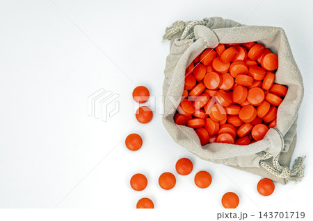 Flat lay top view of open beige fabric drawstring pouch filled with bright orange round medicine tablets, several pills scattered on clean white background, pharmaceutical healthcare and mystery 143701719