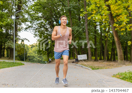Young man smiling while running with blue headphones and gray sneakers on paved walkway in city park Young man smiling while running with blue headphones and gray sneakers on paved walkway in city park 143704467