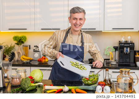 Middle-aged man preparing fresh vegetable salad in kitchen for healthy lunch and home food routine 143704491