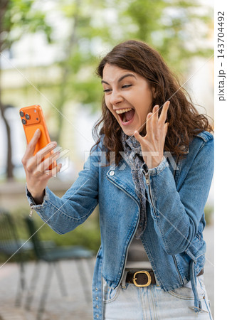 Young woman celebrating smartphone news walking in city street smiling happy in urban park outdoors 143704492