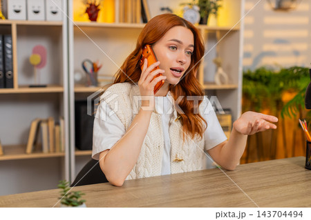 Young woman on home table talking on smartphone with expressive reaction during phone call indoors Young woman on home table talking on smartphone with expressive reaction during phone call indoors 143704494