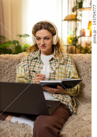 Young woman on home sofa in headphones studies online course on laptop taking careful notes indoors 143704777