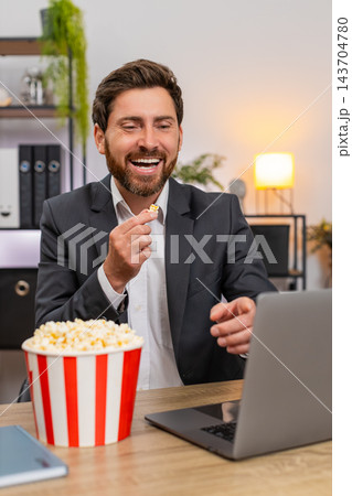 Middle-aged businessman laughs eating popcorn while watching movie on laptop during lunch break 143704780
