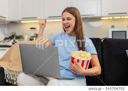 Young woman streams match on laptop celebrates goal claps smiles gives thumbs up eats popcorn snack Young woman streams match on laptop celebrates goal claps smiles gives thumbs up eats popcorn snack 143704782