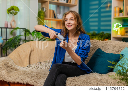 Young woman on home sofa plays online game with gamepad and celebrates success in cozy living room 143704822