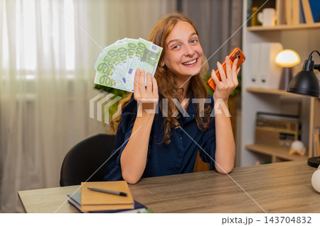 Young woman on home table holding cash and smartphone smiling after online payment success at home Young woman on home table holding cash and smartphone smiling after online payment success at home 143704832