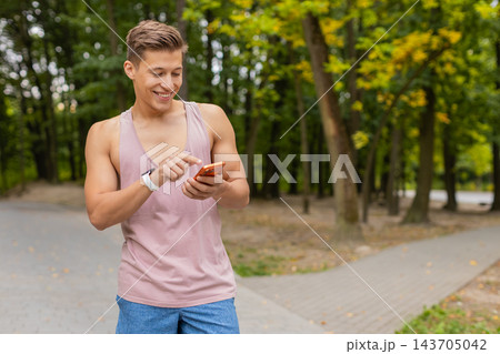 Young man smiling down at orange smartphone while wearing pink tank top and smartwatch in city park 143705042