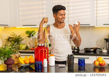 Indian man approving sports supplement with measuring tape for fitness diet progress in home kitchen 143705043