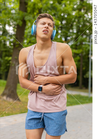 Young man holding stomach with a pained expression and headphones in city park during fitness break Young man holding stomach with a pained expression and headphones in city park during fitness break 143705047