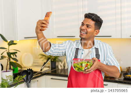 Indian man showing vegetable salad to smartphone while sharing healthy meal tips in modern kitchen 143705065