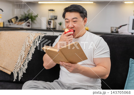 Asian man bites red apple enjoys juicy taste and read short health tips in book with calm smile well 143705068