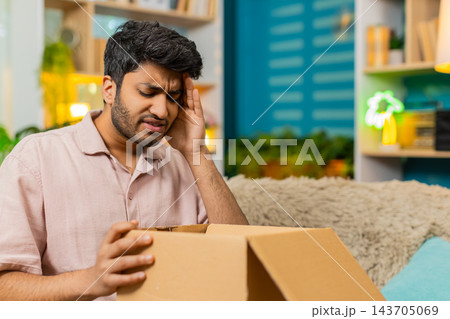 Indian man on home sofa opening parcel finding wrong product with upset face during online shopping Indian man on home sofa opening parcel finding wrong product with upset face during online shopping 143705069
