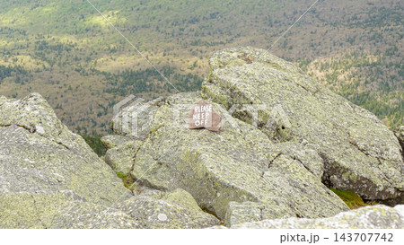 Weathered please keep off sign on lichen covered granite rocks overlooking a forested mountain landscape from a rugged summit Weathered please keep off sign on lichen covered granite rocks overlooking a forested mountain landscape from a rugged summit 143707742