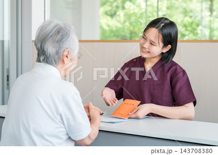 介護施設で年金手帳などの書類を確認するシニア女性と介護スタッフ。老人ホームの入居手続き 介護施設で年金手帳などの書類を確認するシニア女性と介護スタッフ。老人ホームの入居手続き 143708380
