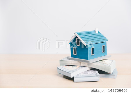 A blue model house rests on several stacks of banknotes against a light background with open space on the left, illustrating home purchase costs, housing affordability, property financing concepts A blue model house rests on several stacks of banknotes against a light background with open space on the left, illustrating home purchase costs, housing affordability, property financing concepts 143709493