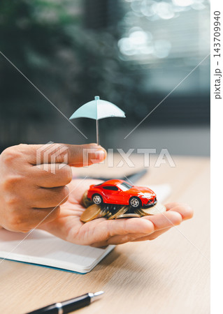 A red toy car rests on coins in a person's palm beneath a small blue umbrella, with a notebook and pen on the desk, illustrating vehicle savings and financial protection as a concept. 143709940