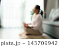 A softly blurred person sits cross-legged on the floor near a bright window, with a sofa beside them and open space on the left, creating a gentle indoor backdrop for wellness-themed design. 143709942