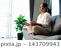 A woman sits cross-legged on a gray sofa with both hands forming meditation-style gestures, beside a leafy potted plant and bright window, leaving open space on the left for wellness messaging. 143709943