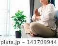 A woman sits cross-legged on a gray sofa with eyes closed and palms pressed together at chest level, beside a potted plant and bright window, leaving space on the left for wellness text. 143709944