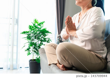 A woman sits cross-legged on a gray sofa with eyes closed and palms pressed together at chest level, beside a potted plant and bright window, leaving space on the left for wellness text. 143709944