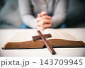 A wooden cross lies across the center of an open book on a white table, while a seated person's clasped hands appear softly blurred behind it, creating a horizontal Christian faith image. 143709945