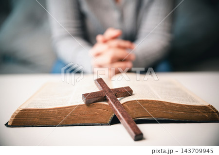 A wooden cross lies across the center of an open book on a white table, while a seated person's clasped hands appear softly blurred behind it, creating a horizontal Christian faith image. 143709945