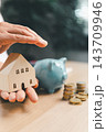 A person holds a wooden model house in one palm while another hand hovers above it, with a blue piggy bank and stacks of coins nearby, illustrating home savings and housing budget planning. 143709946