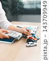A person holds a stack of banknotes beside a miniature blue umbrella positioned above four toy cars, creating a vertical composition about vehicle-related financial planning. 143709949