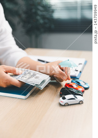 A person holds a stack of banknotes beside a miniature blue umbrella positioned above four toy cars, creating a vertical composition about vehicle-related financial planning. A person holds a stack of banknotes beside a miniature blue umbrella positioned above four toy cars, creating a vertical composition about vehicle-related financial planning. 143709949