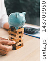 A light blue piggy bank stands on a wooden block tower as a finger touches a lower block, with a calculator blurred in the background, suggesting savings and financial stability as a concept. 143709950
