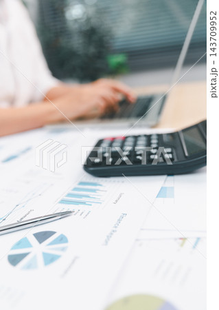 Financial report with charts, pen, and calculator on an office desk while a professional uses a laptop, representing accounting, budgeting, analytics, finance, planning, and business research. Financial report with charts, pen, and calculator on an office desk while a professional uses a laptop, representing accounting, budgeting, analytics, finance, planning, and business research. 143709952