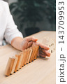 A hand is positioned between a diagonal row of leaning wooden domino blocks and a model house, illustrating a protective barrier against a potential domino effect in a vertical composition. 143709953