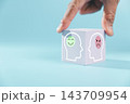 A hand holds a light-colored cube showing a smiling face inside one head outline and a frowning face inside another, creating a visual contrast between emotional expressions on a blue background. 143709954