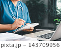 Healthcare professional in blue scrubs writing notes in a small notebook beside a laptop, representing clinical documentation, patient planning, medical studies, and daily healthcare administration. 143709956