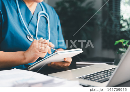 Healthcare professional in blue scrubs writing notes in a small notebook beside a laptop, representing clinical documentation, patient planning, medical studies, and daily healthcare administration. Healthcare professional in blue scrubs writing notes in a small notebook beside a laptop, representing clinical documentation, patient planning, medical studies, and daily healthcare administration. 143709956